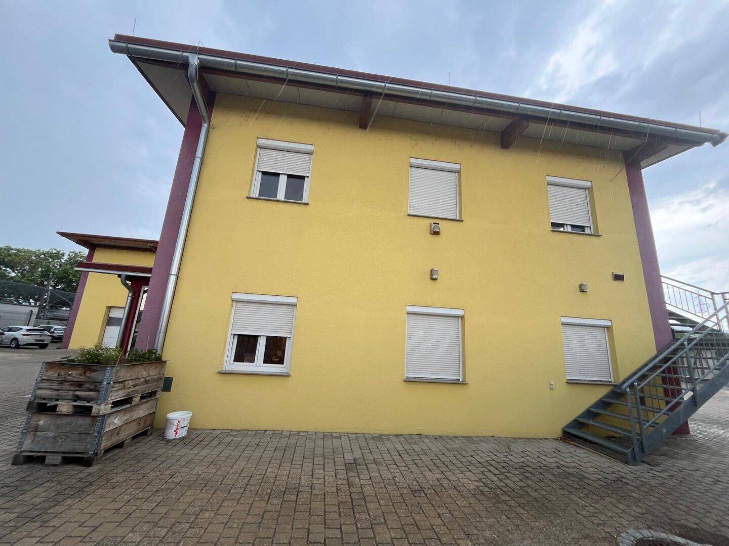 Gelbes Gebäude mit mehreren Fenstern und Rollläden, dazu eine Außentreppe und gepflasterter Vorplatz.