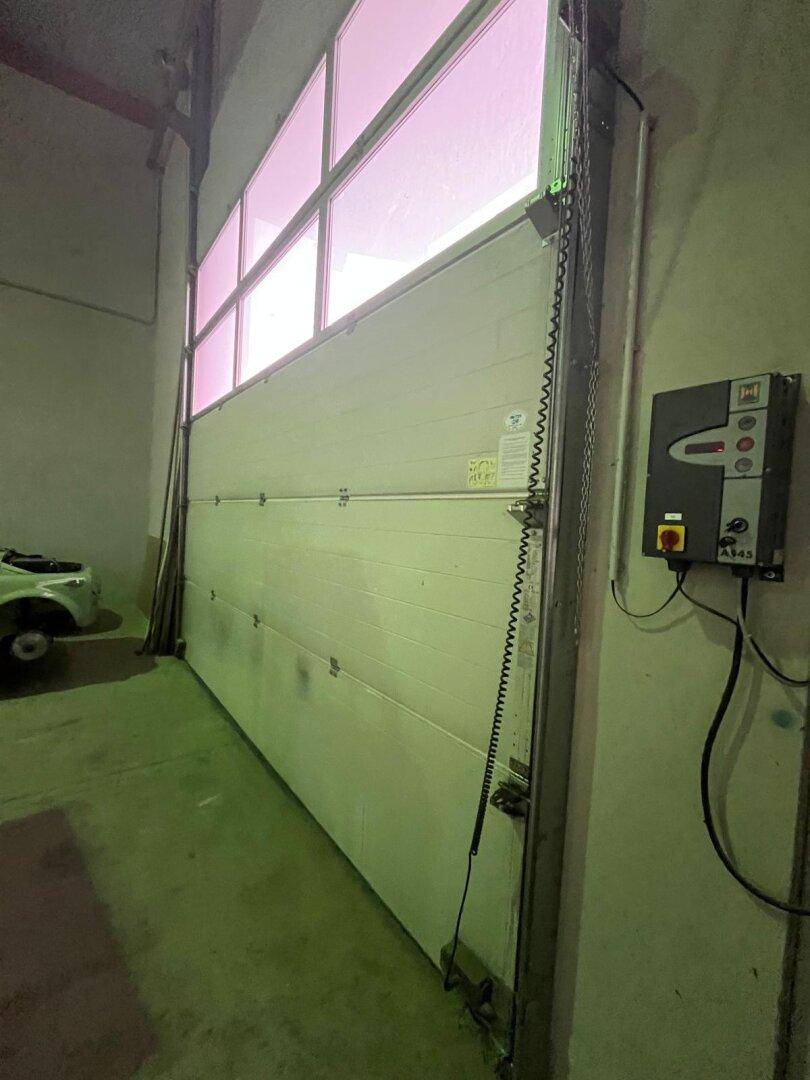 Großes weißes Industrierolltor mit Fenstern und einem elektrischen Bedienfeld an der Wand montiert.