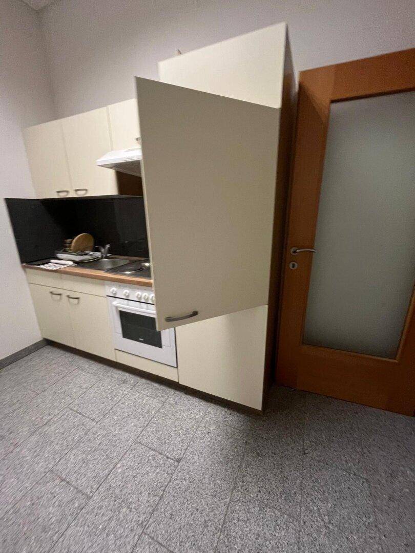 Kleine Küchenzeile mit beigen Schränken, Herd und Backofen, daneben eine Holztür mit Milchglas.
