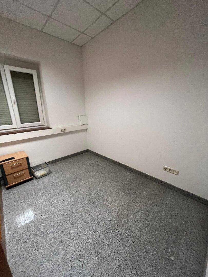 Leeres Büro mit grauem Fliesenboden, weiß gestrichenen Wänden und einem Fenster mit Jalousien.