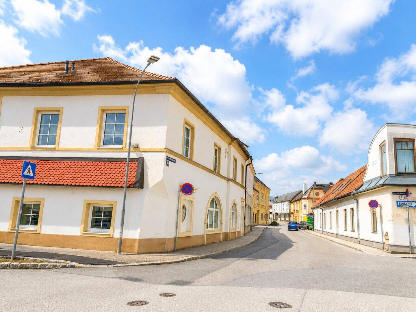 Historisches Gebäude an einer Straßenecke mit weißer und gelber Fassade sowie rotem Ziegeldach unter blauem Himmel.