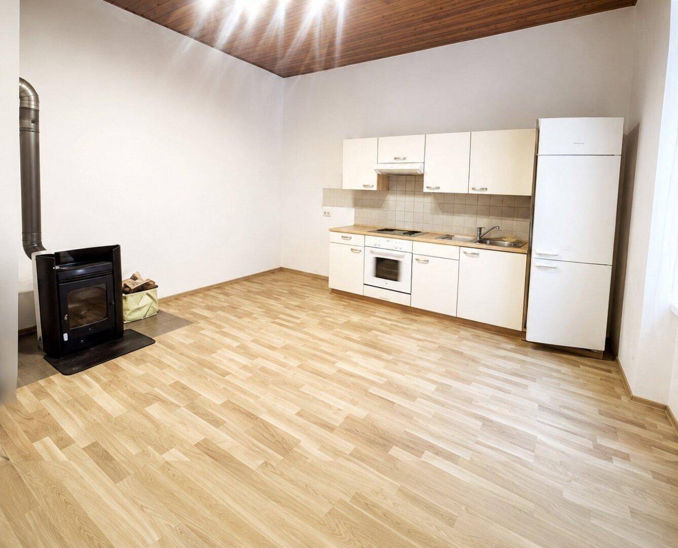 Wohnküche mit Holzofen, Einbauküche, Holzdecke und Laminatboden, gut beleuchtet.