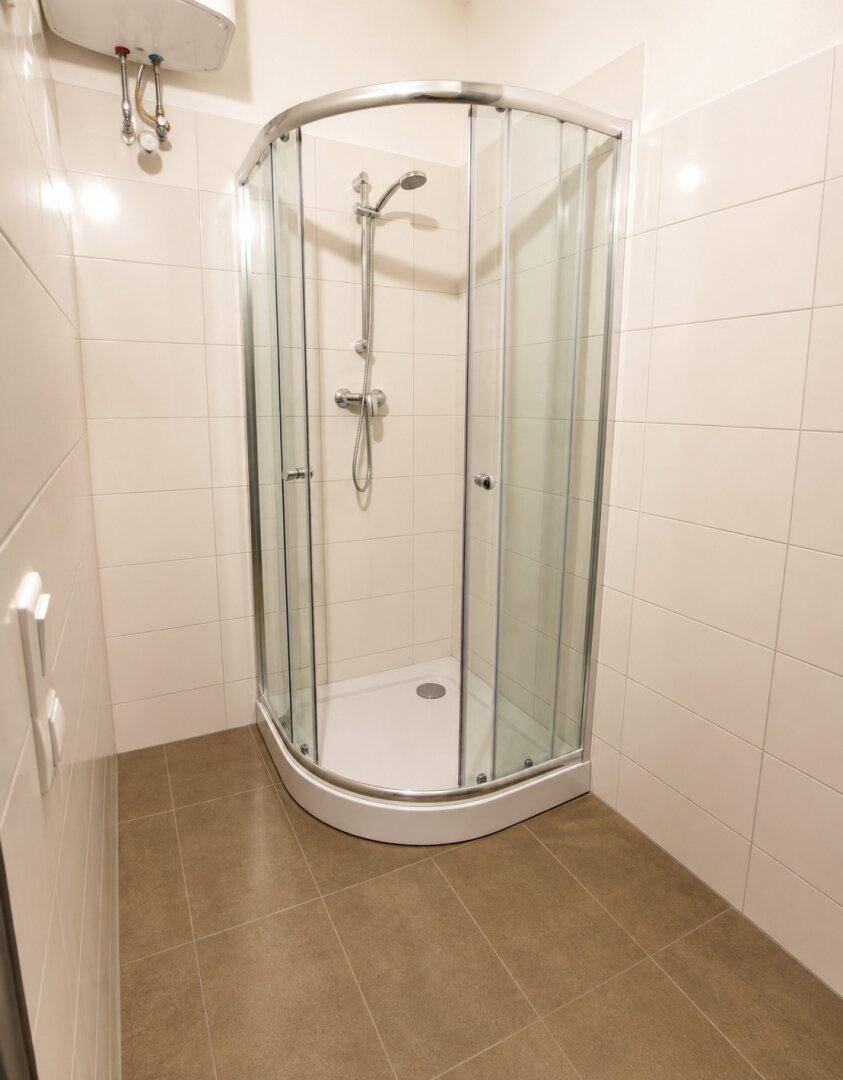 Badezimmer mit Eckduschkabine, weißen Wandfliesen und grauen Bodenfliesen, inklusive Warmwasserbereiter.