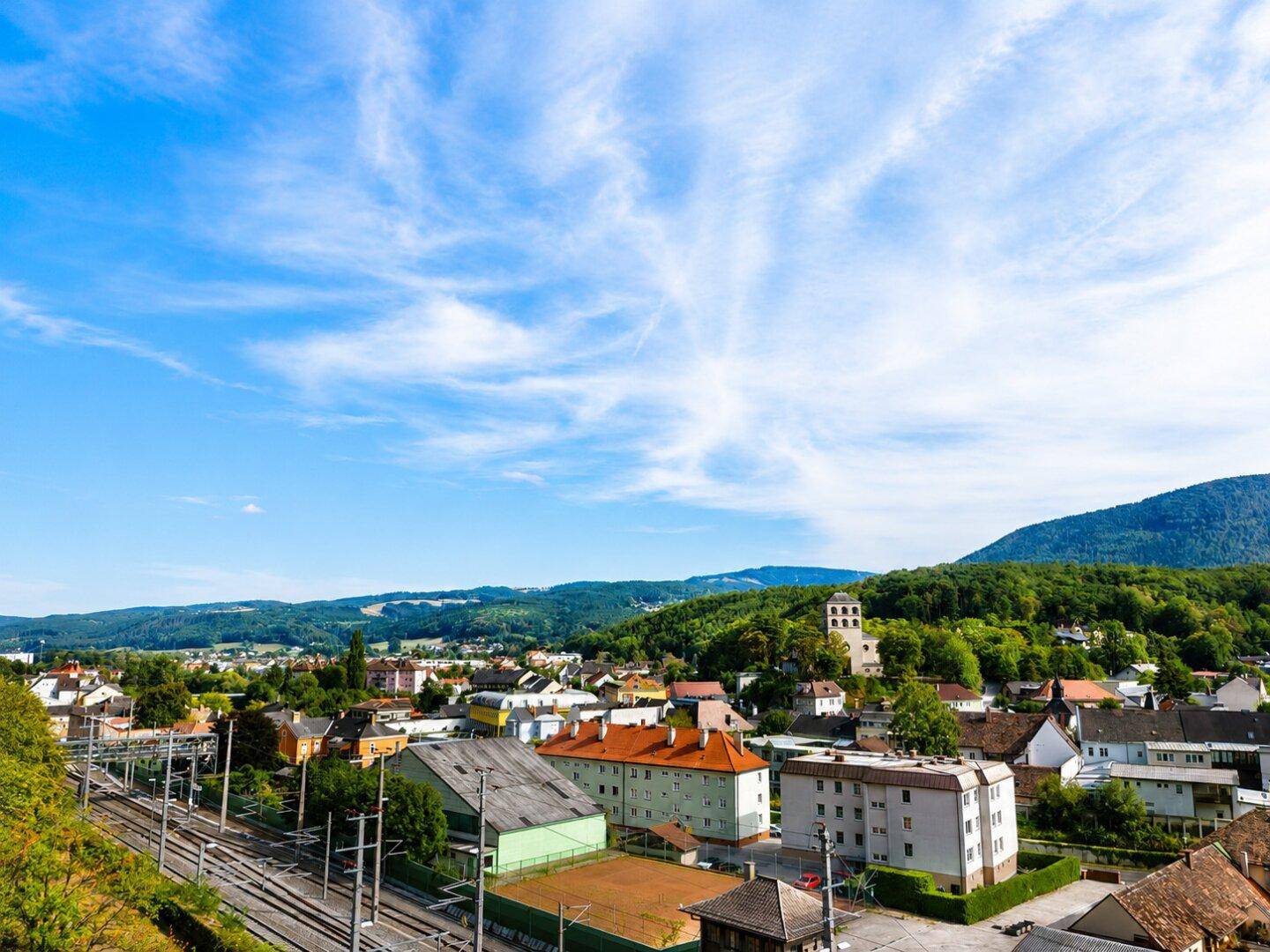 Weitläufiger Blick auf eine Stadt mit Bahngleisen, umgeben von bewaldeten Bergen unter einem klaren Himmel.