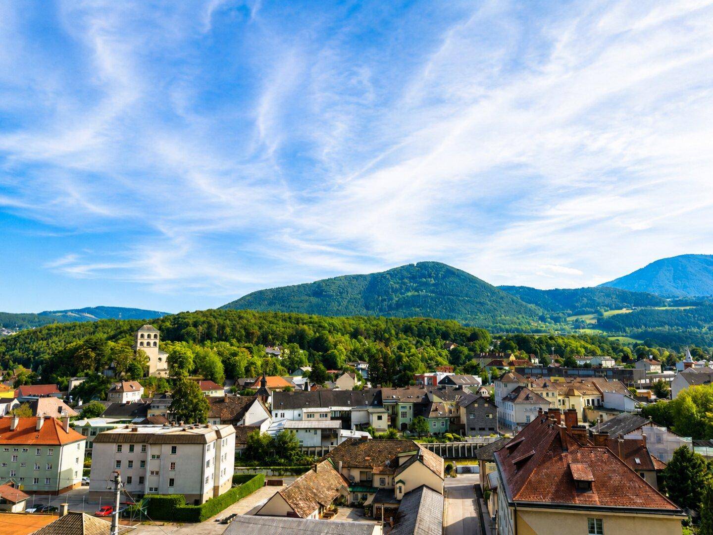 Panoramablick über eine malerische Stadt mit grünen Hügeln und Bergen im Hintergrund unter blauem Himmel.