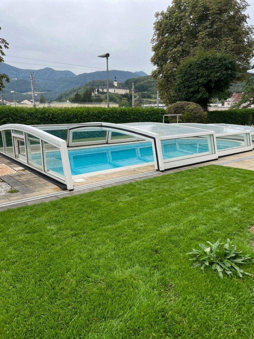 Außenpool mit verschiebbarer Überdachung und Rasenfläche im Gartenbereich, umgeben von grünen Hügeln.