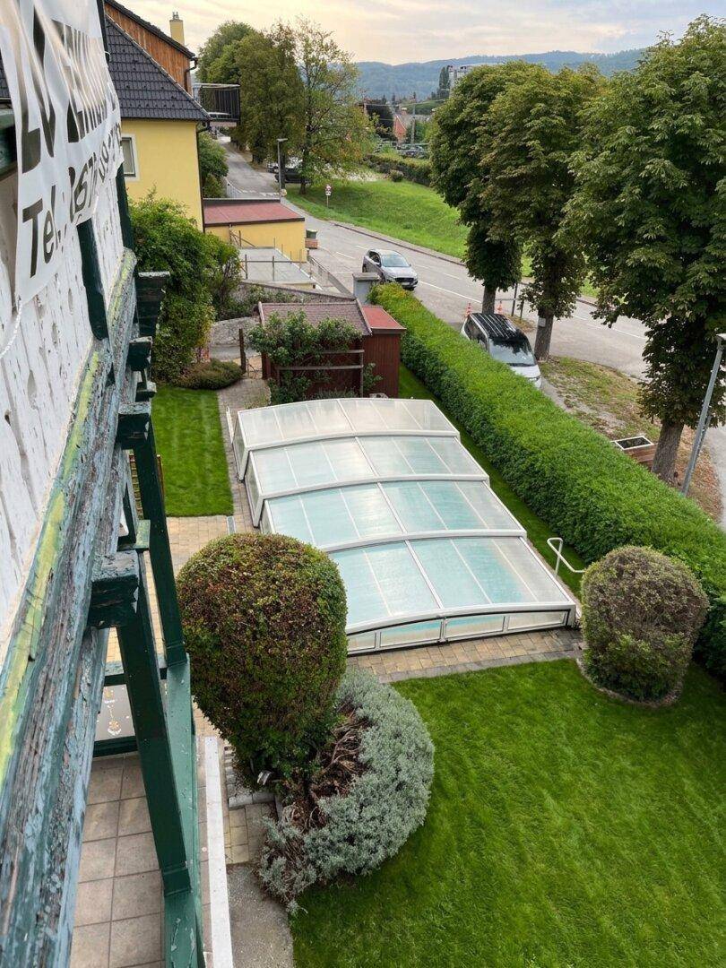 Blick von der Loggia auf den gepflegten Garten mit überdachtem Pool und Straße.
