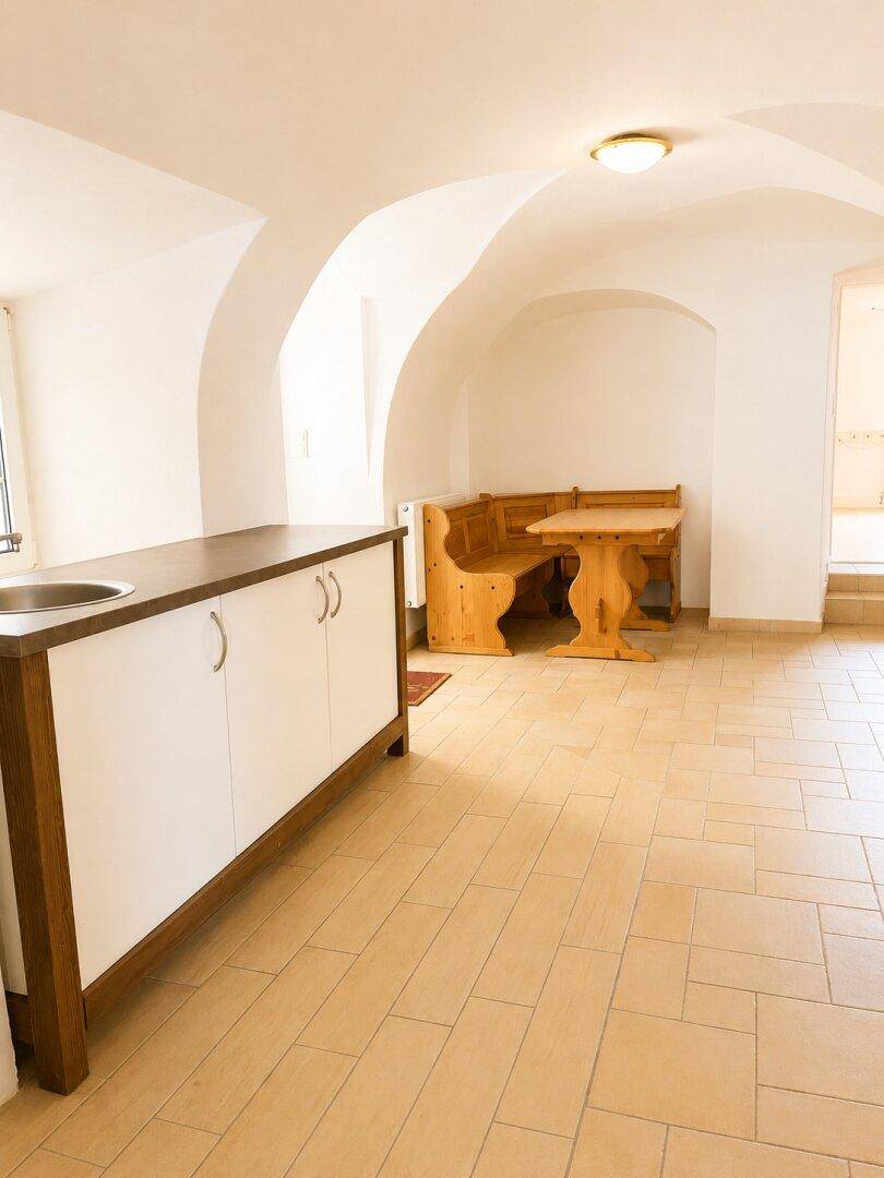 Raum mit Gewölbedecke, Fliesenboden, kleiner Küchenzeile und rustikaler Sitzecke aus Holz.