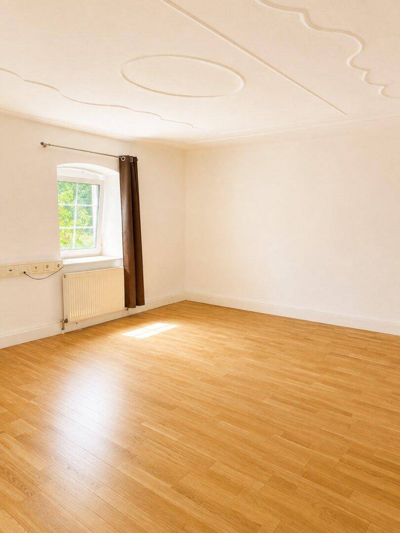 Heller Raum mit Holzparkett, weißen Wänden, einem Fenster und Heizkörper unterhalb, gut beleuchtet.