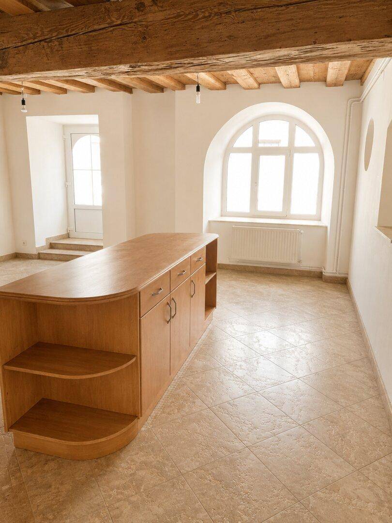 Heller Raum mit Holzbalkendecke, Fliesenboden und einer Holzinsel, beleuchtet durch ein großes Rundbogenfenster.
