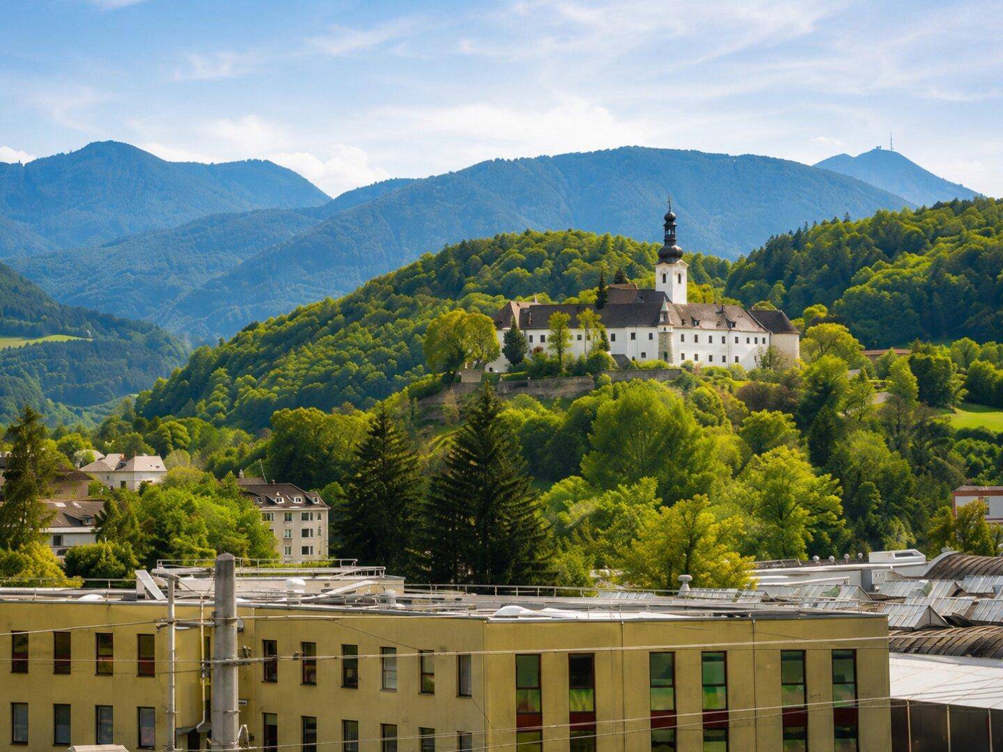 Historisches Klostergebäude auf einem bewaldeten Hügel, umgeben von einer beeindruckenden Berglandschaft.
