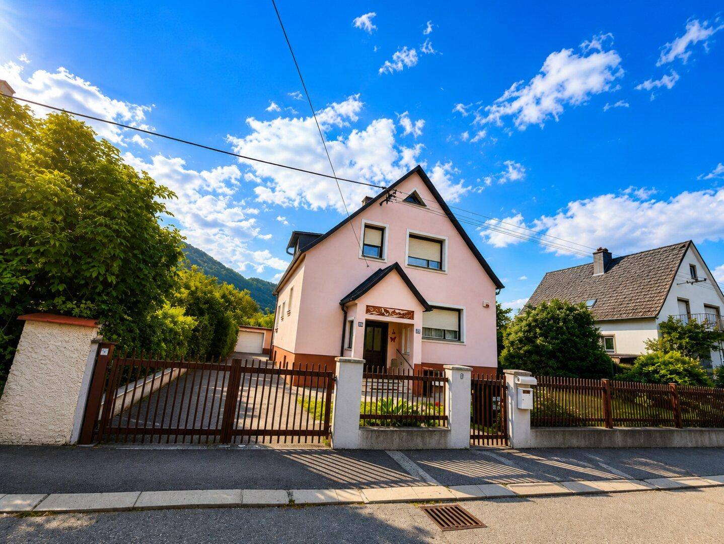 Rosa Hausfassade mit Zaun und Einfahrt, umgeben von grüner Landschaft und Bergen.