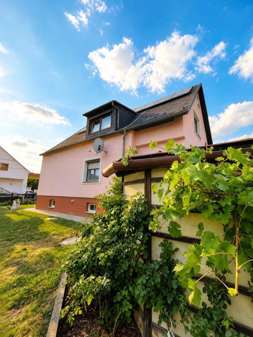 Seitenansicht des Hauses mit Dachschrägen und Photovoltaikanlage unter blauem Himmel.