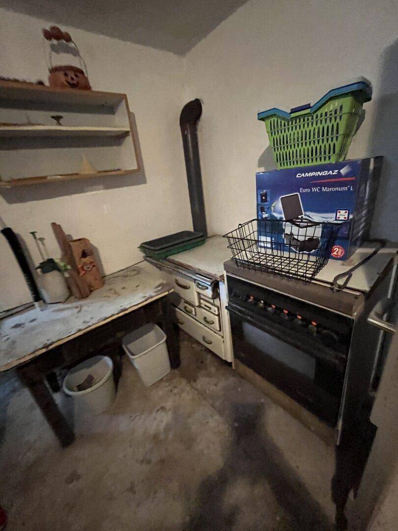 Unaufgeräumter Kellerraum mit altem Ofen, Tisch und verschiedenen Haushaltsgegenständen.