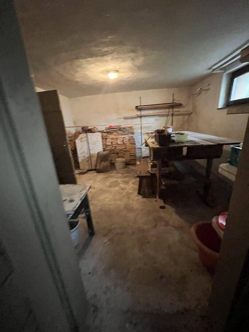 Kellerraum mit Tisch, Regalen und verschiedenen Gegenständen, der als Abstellfläche dient.