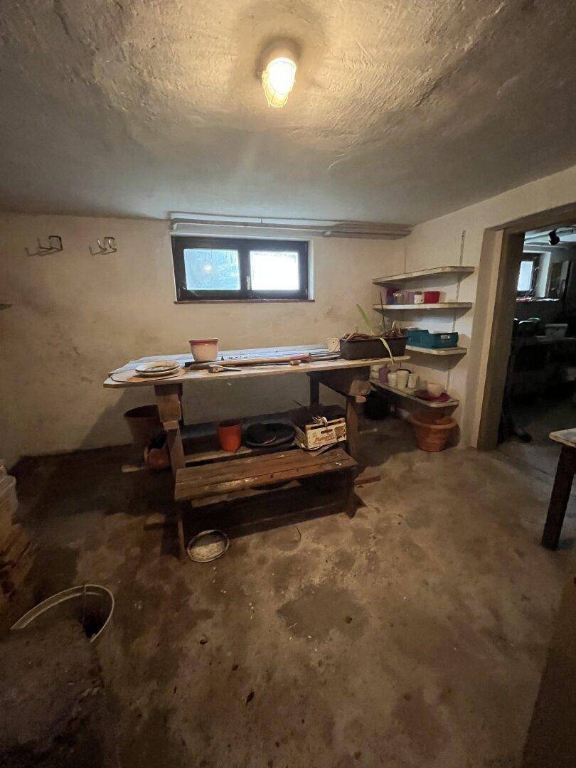 Kellerraum mit einem Arbeitstisch, Regalen und einem Fenster, das Tageslicht hereinlässt.