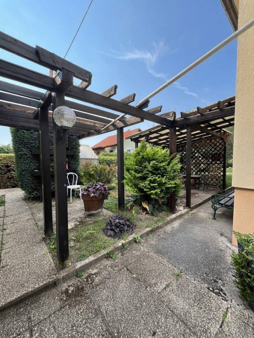 Gartenweg unter einer Holzpergola, umgeben von üppigem Grün und verschiedenen Pflanzen.