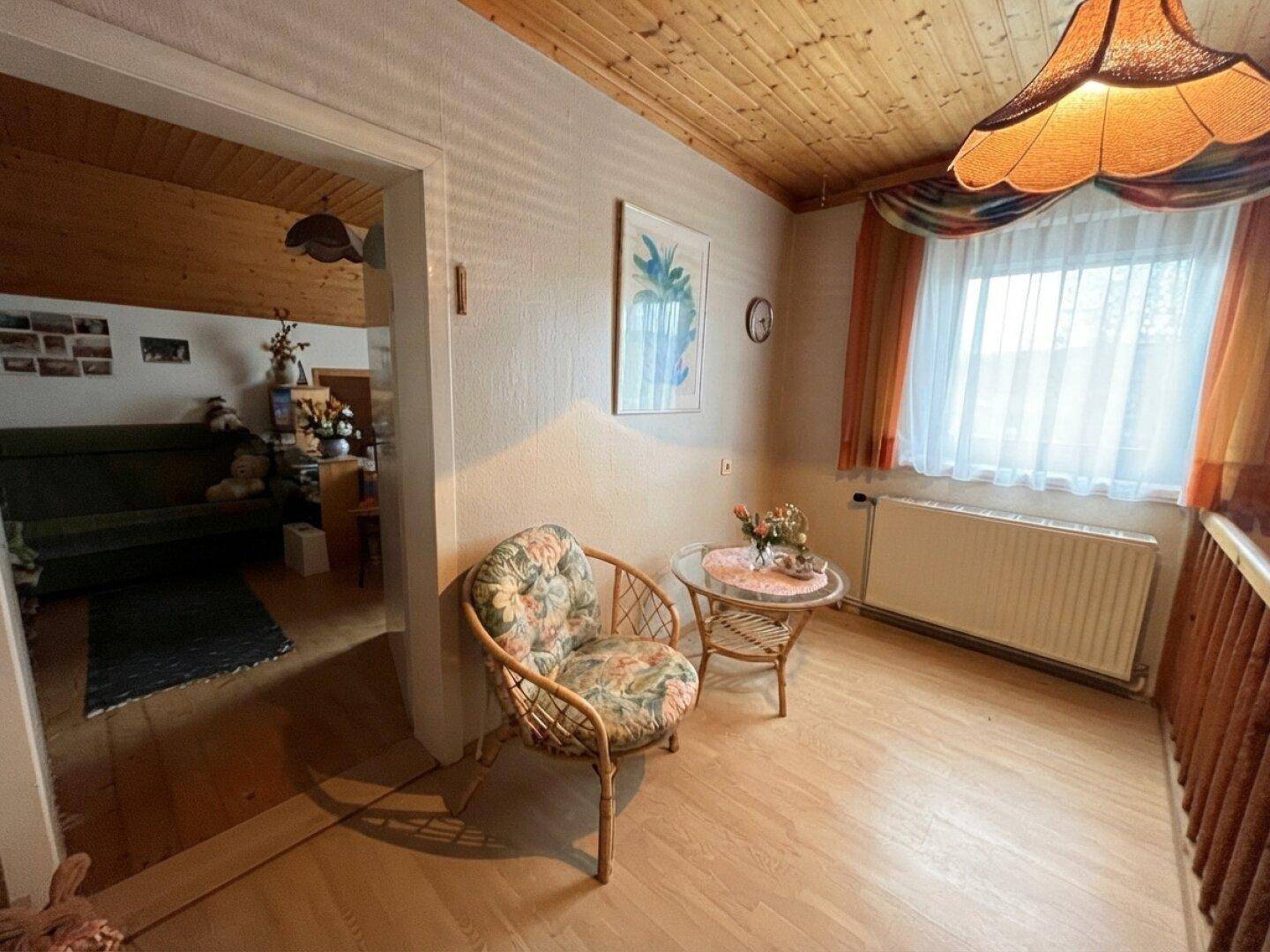 Kleiner Raum mit Korbstuhl und Tisch, Holzboden und Fenster, ideal als Leseecke.