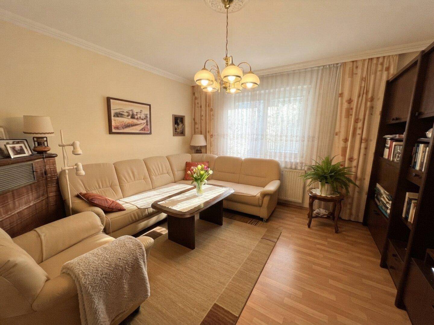 Helles Wohnzimmer mit beigem Sofa, Holzmöbeln und einem Fenster mit Vorhängen.