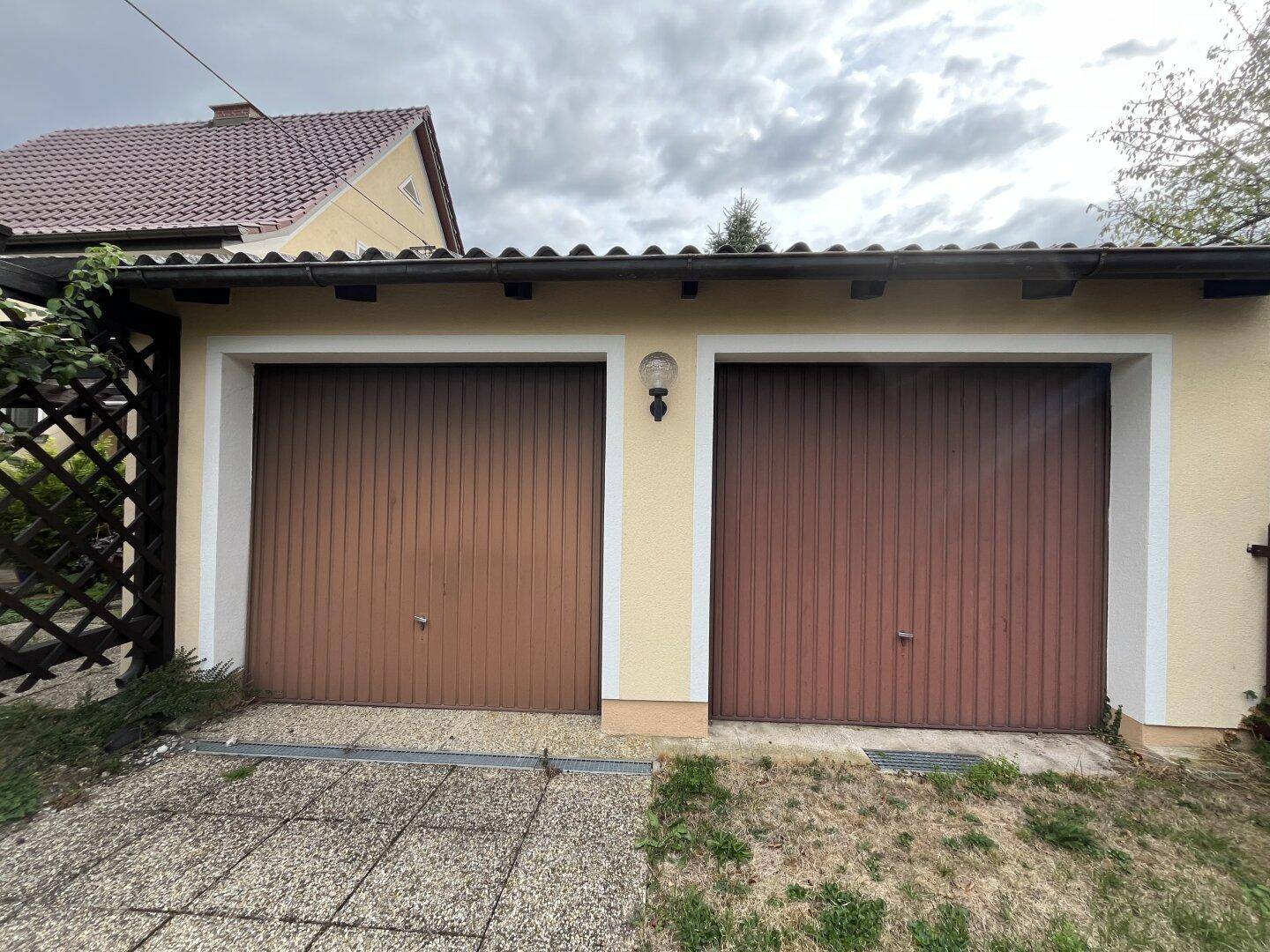 Doppelgarage mit zwei braunen Holztoren und einem kleinen Vorplatz unter bewölktem Himmel.