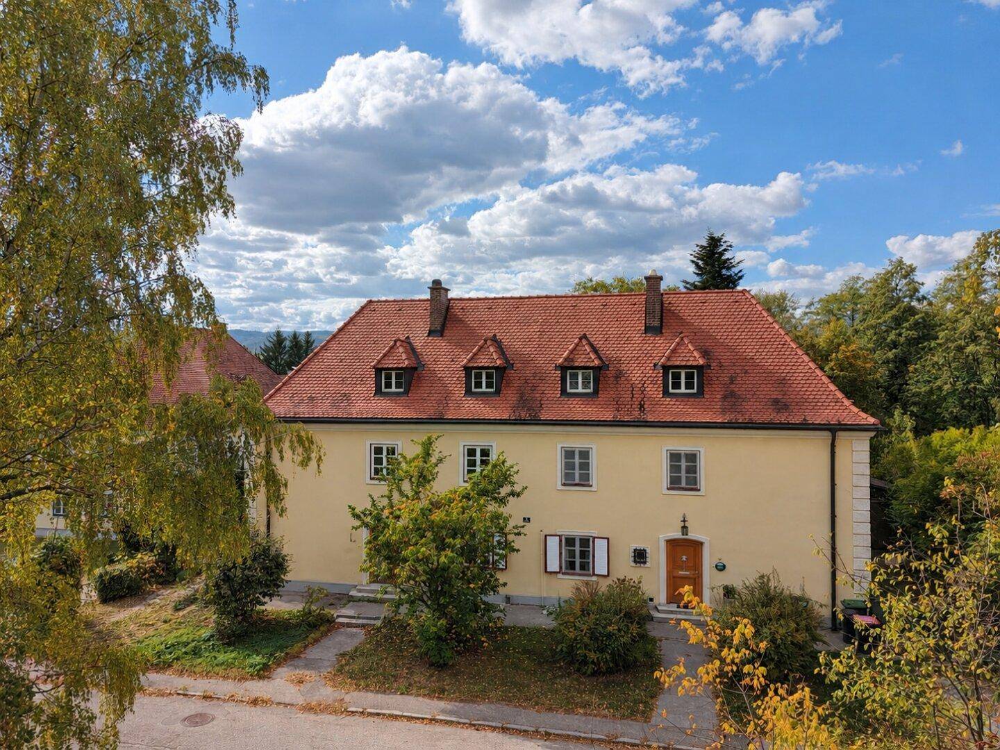 Historisches gelbes Gebäude mit rotem Ziegeldach, umgeben von üppiger Herbstvegetation unter blauem Himmel.