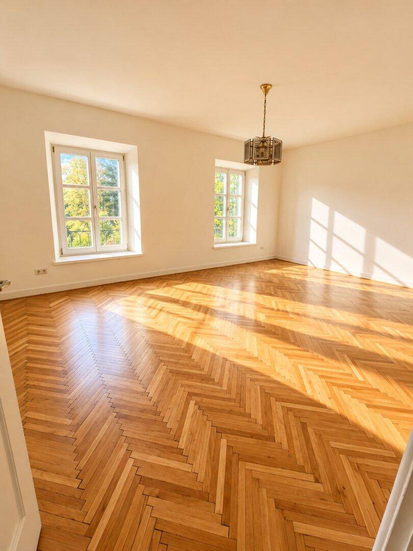 Helles, leeres Zimmer mit Fischgrätparkettboden und zwei großen Fenstern, durchflutet von Sonnenlicht.