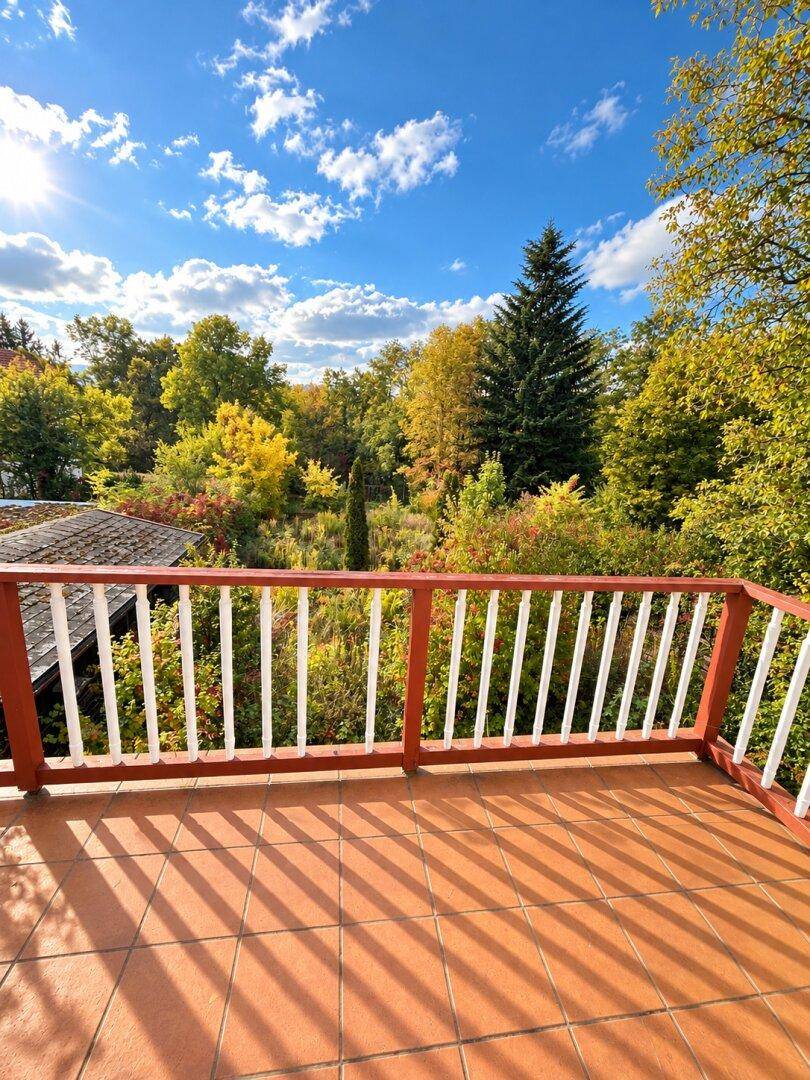 Sonniger Balkon mit Terrakotta-Fliesen und rotem Geländer, umgeben von grüner Natur.