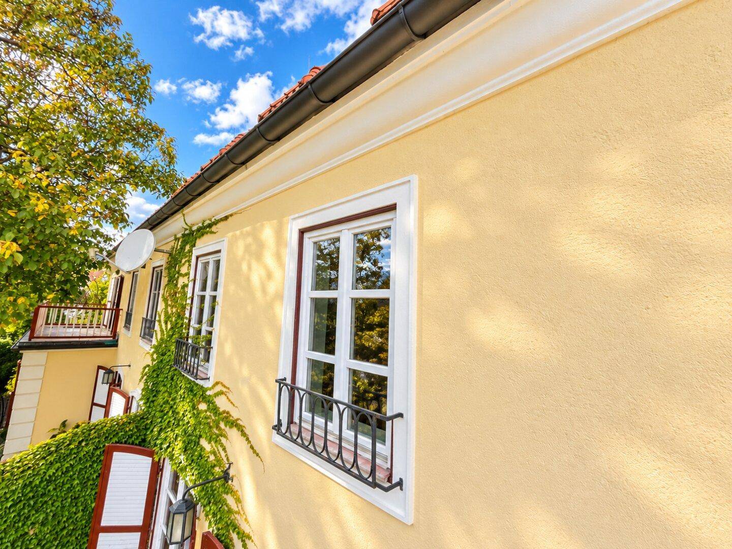 Nahaufnahme der gelben Hausfassade mit weißen Sprossenfenstern und Kletterpflanzen.