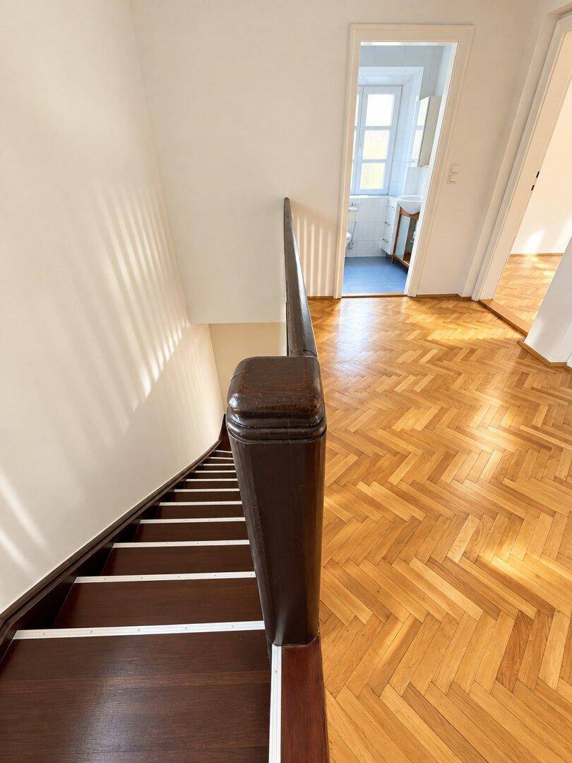 Treppenhaus mit dunklem Holzgeländer und Blick auf einen Raum mit Fischgrätparkett.