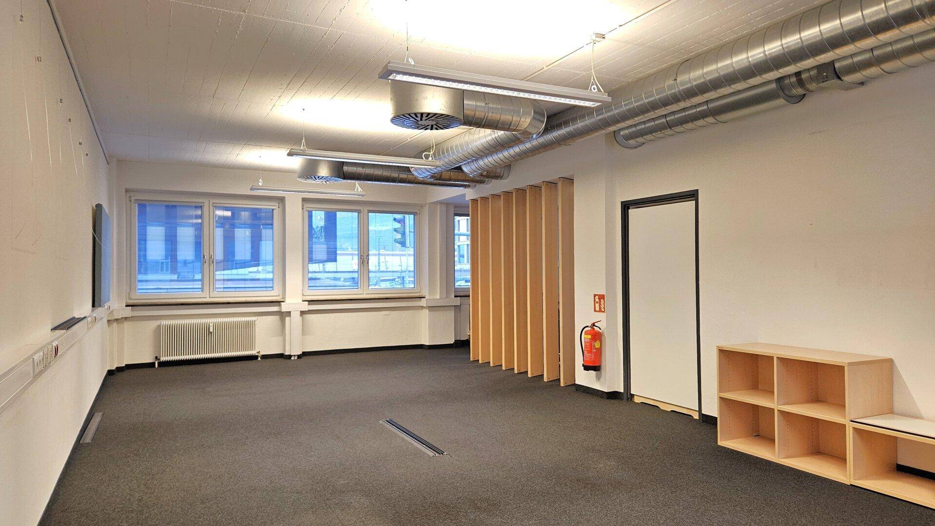 Geräumiger, leerer Büroraum mit Teppichboden, großen Fenstern und sichtbaren Lüftungsrohren an der Decke.