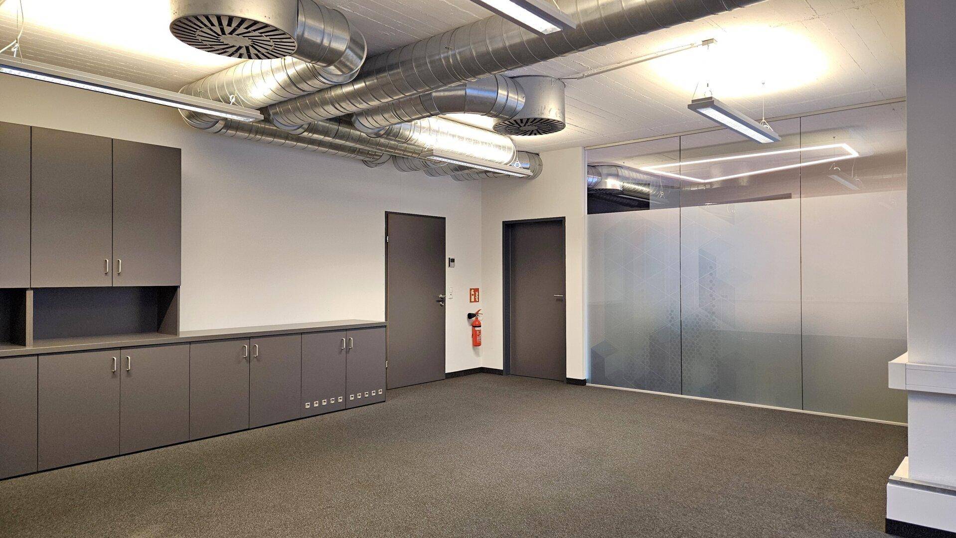 Heller Büroraum mit grauen Einbauschränken, Teppichboden und sichtbaren Lüftungsrohren an der Decke.