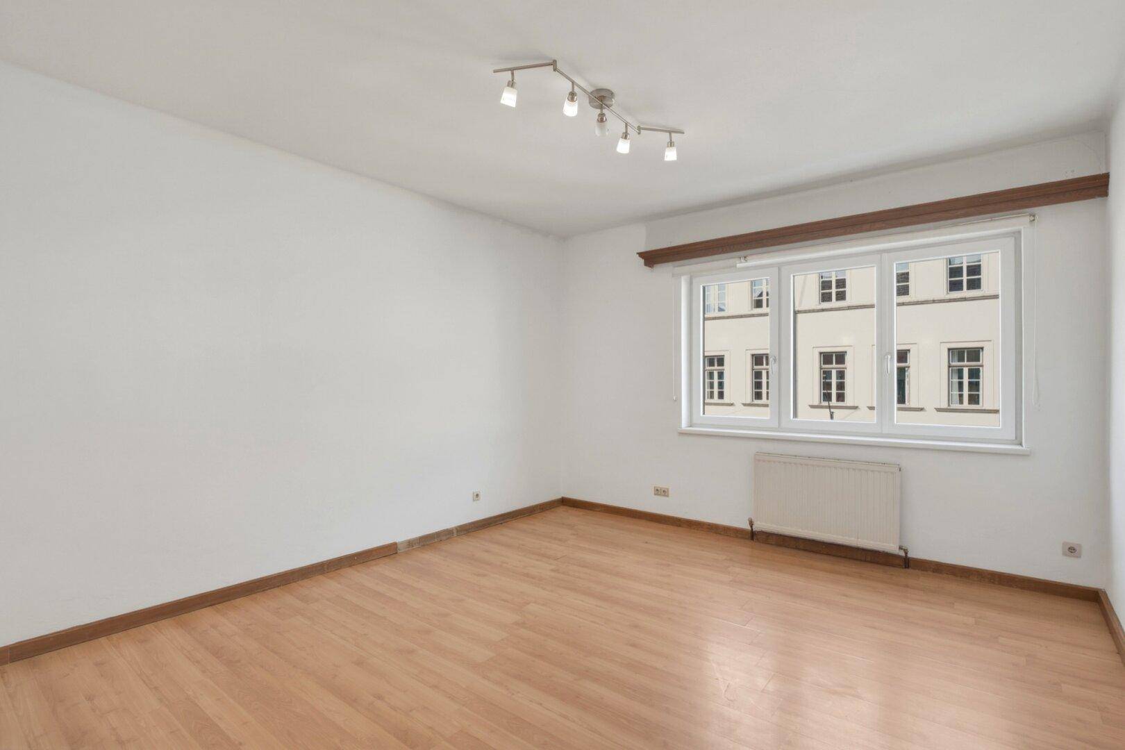 Leeres Wohnzimmer mit hellem Laminatboden, weißen Wänden und einem Fenster mit Blick auf die Stadt.