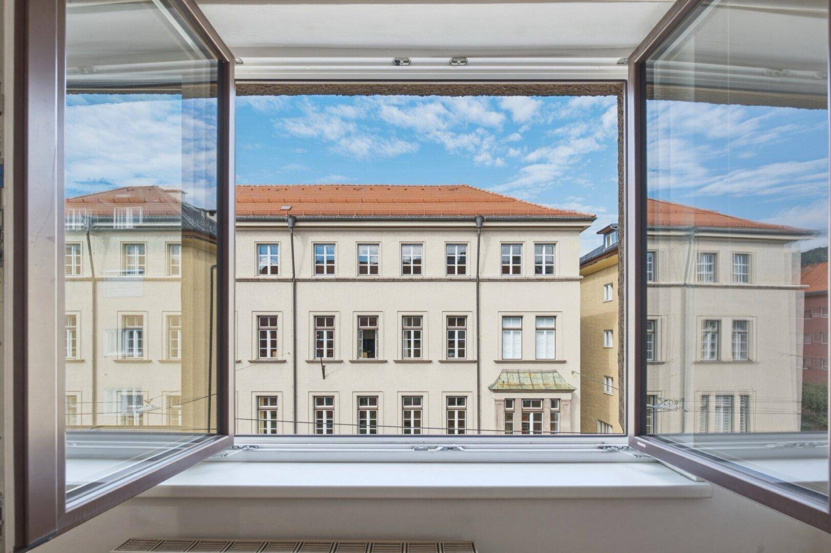 Blick aus einem geöffneten Fenster auf die Fassade eines gegenüberliegenden historischen Gebäudes unter blauem Himmel.