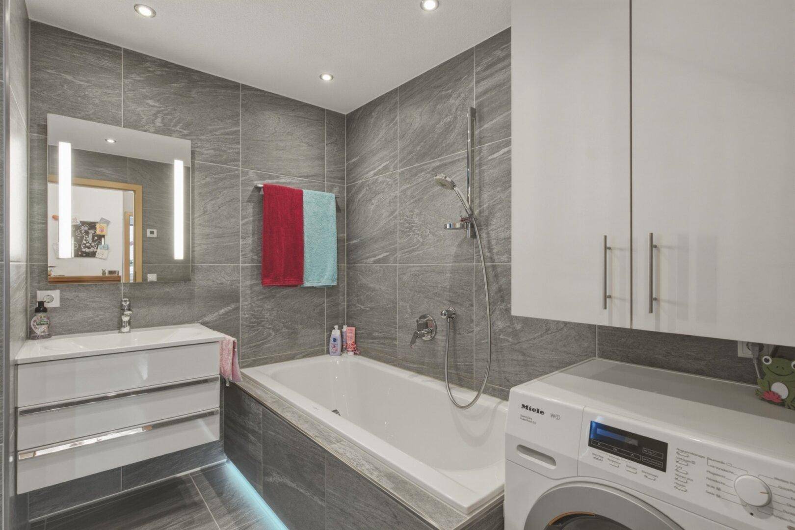 Modernes Badezimmer mit grauen Fliesen, Badewanne mit Duschfunktion und Waschmaschine.