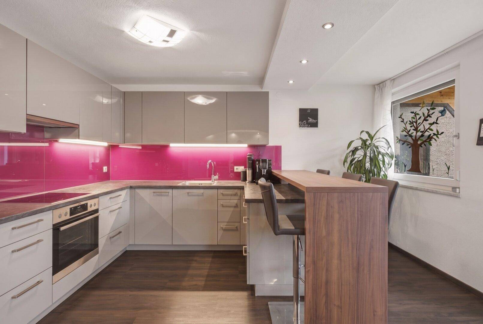 Moderne Küche mit grauen Hochglanzfronten, pinker Rückwand und integrierter Frühstücksbar.