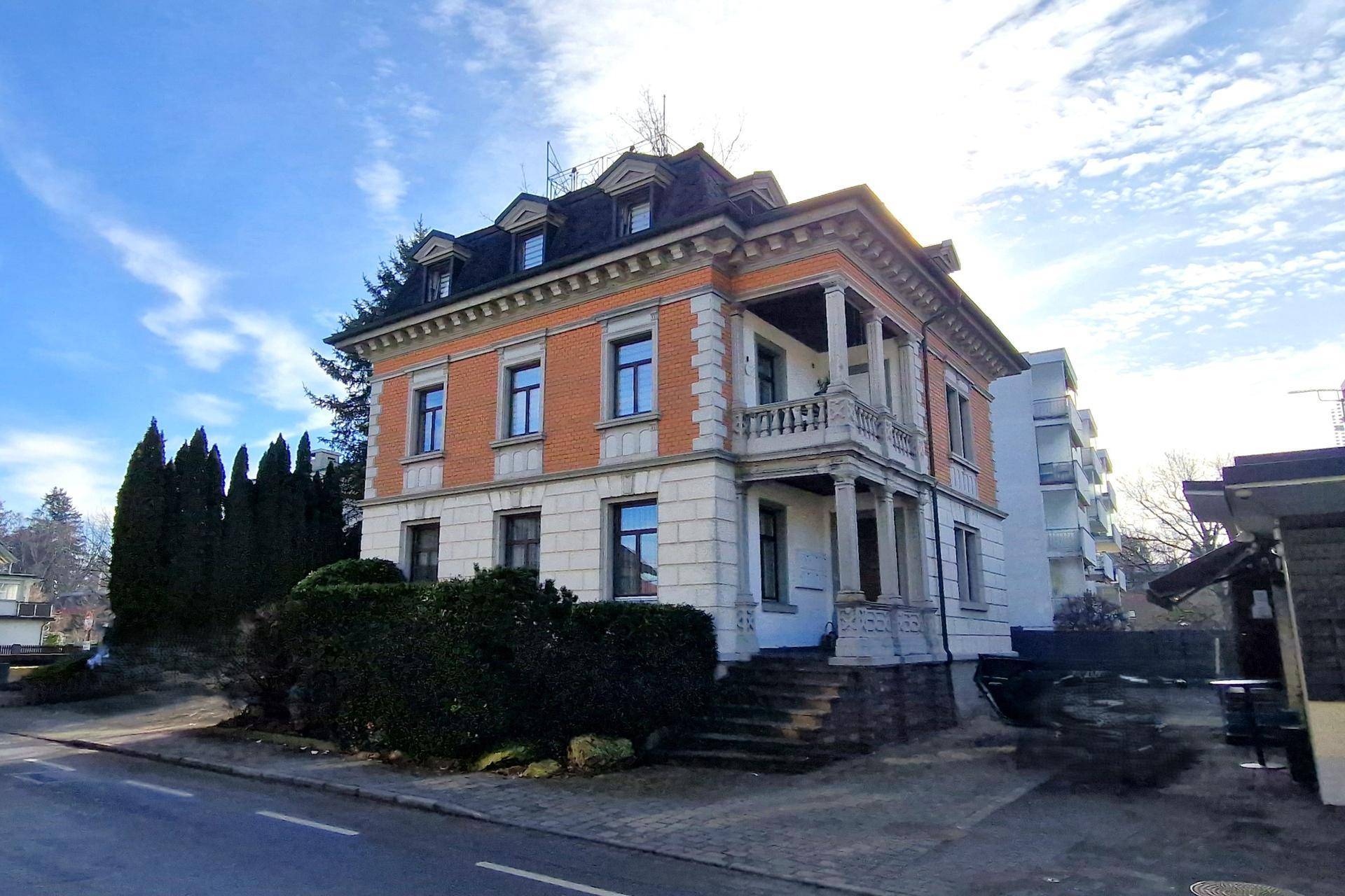 Klassisches Gebäude mit Mansarddach, Balkon und Ziegelfassade unter strahlendem Himmel.