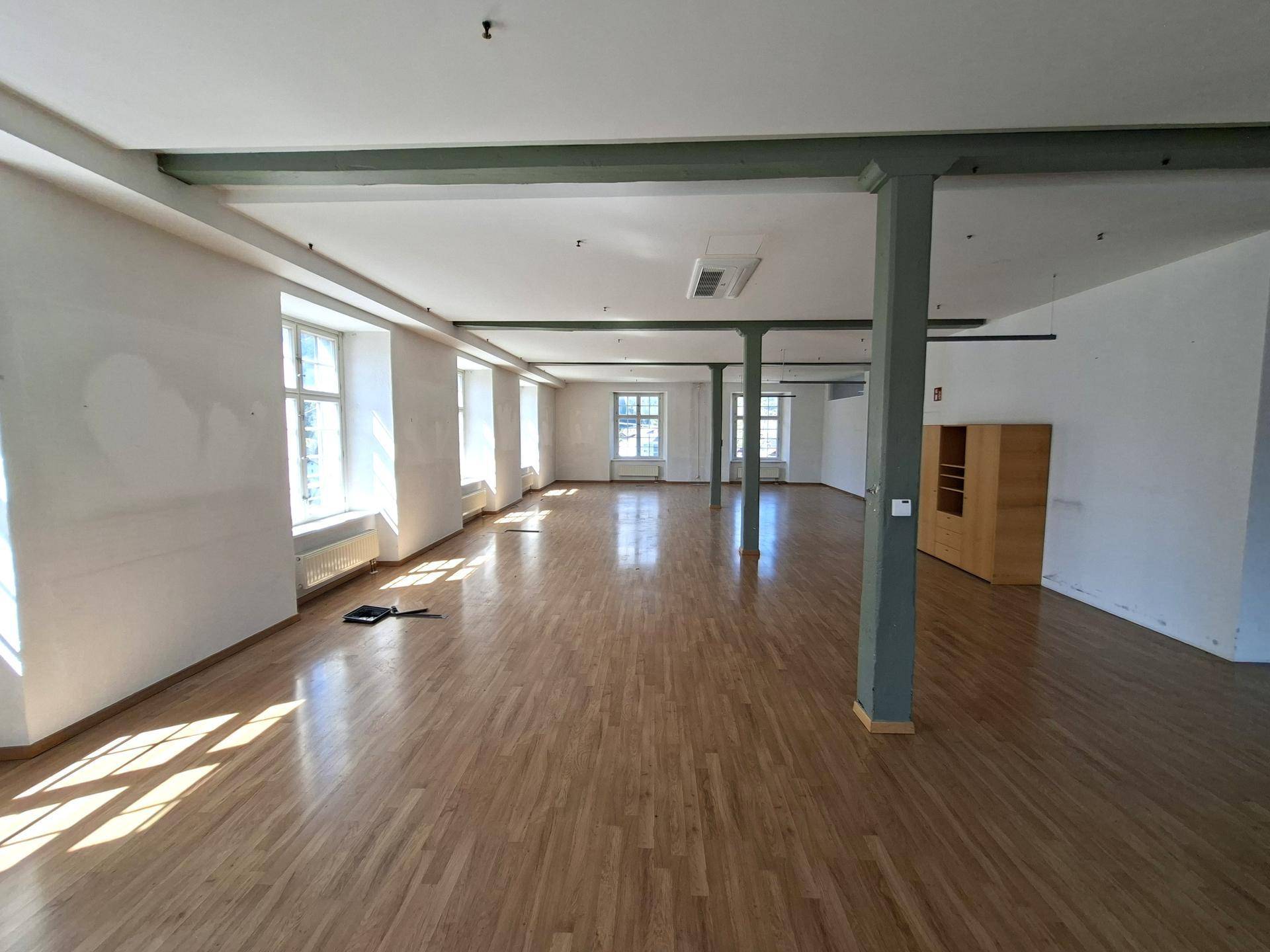 Langer, heller Büroraum mit Holzboden, grünen Stützpfeilern und vielen Fenstern für Tageslicht.