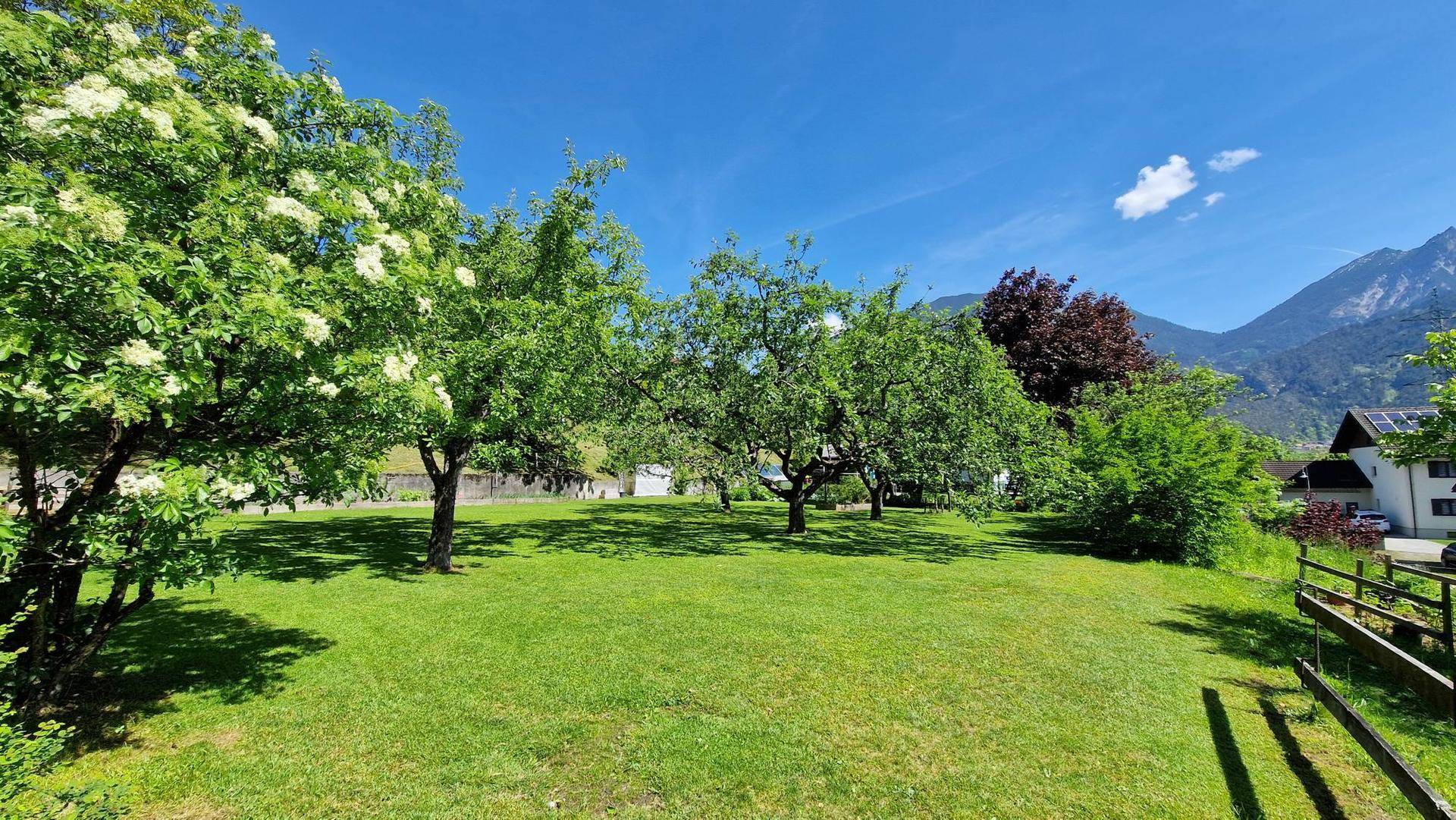 Weitläufiger Garten mit blühenden Bäumen, Rasen und Blick auf die umliegende Berglandschaft unter blauem Himmel.