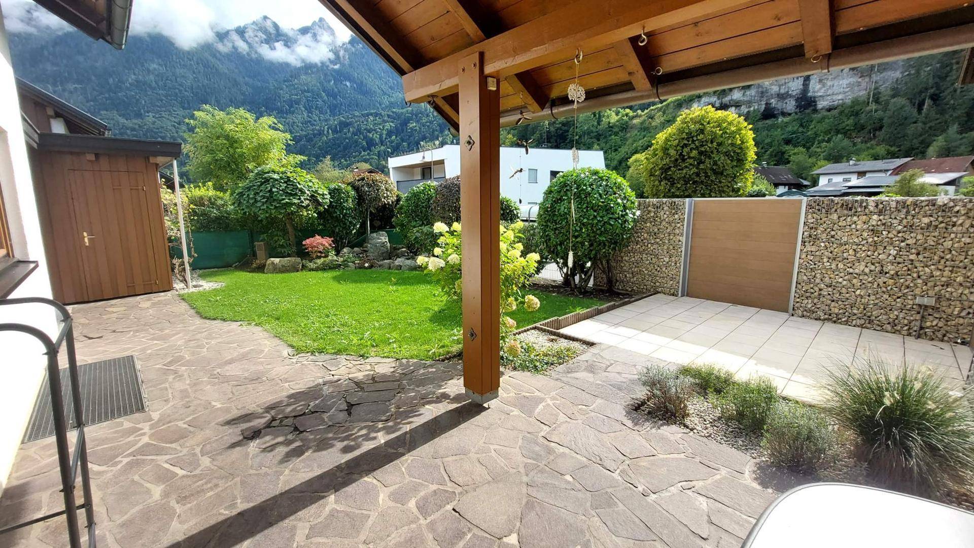Gepflasterte Terrasse mit Rasenfläche, Gartenhaus und einem beeindruckenden Bergblick.
