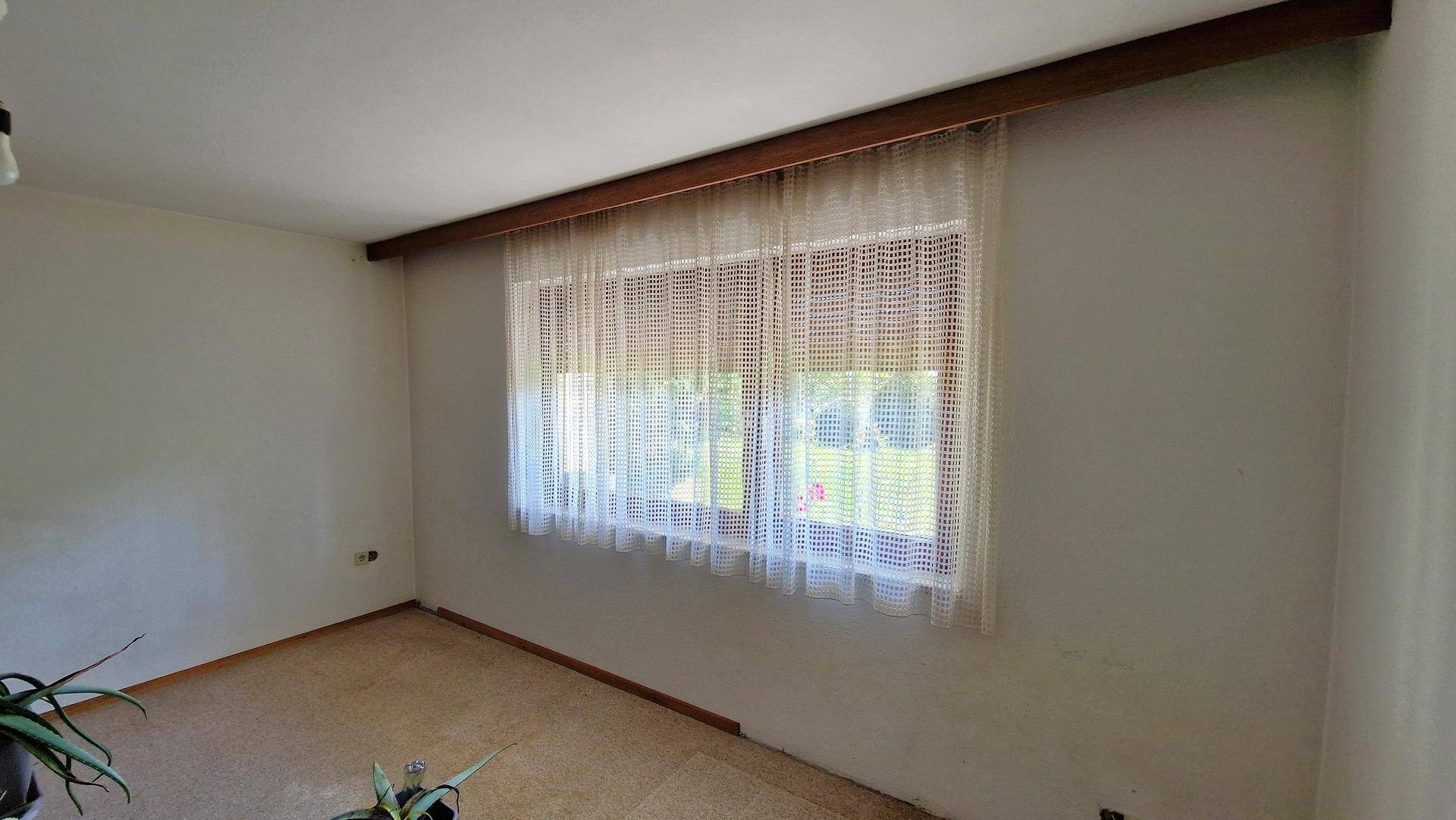 Einfaches Zimmer mit Teppichboden und einem großen Fenster, das viel Tageslicht hereinlässt.