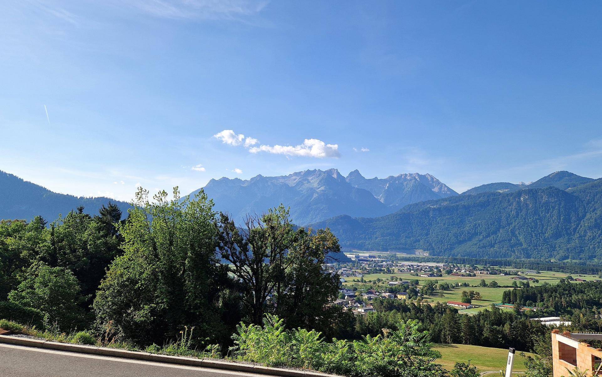 Weitläufiger Blick über ein grünes Tal mit einer Stadt, umgeben von Wäldern und majestätischen Bergen.