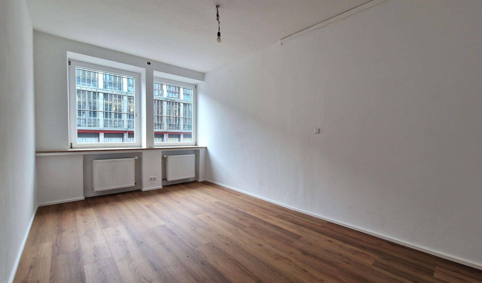 Heller Wohnraum mit Laminatboden, weißen Wänden und zwei großen Fenstern, die für viel Tageslicht sorgen.