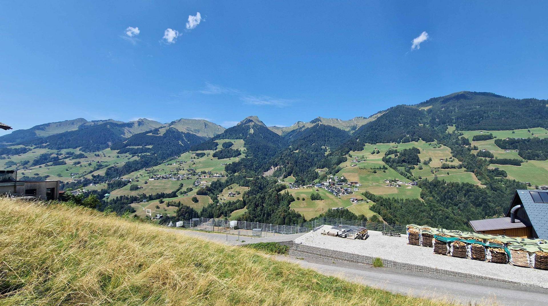 Panoramablick auf eine weite grüne Berglandschaft mit kleinen Dörfern und klarem blauem Himmel.