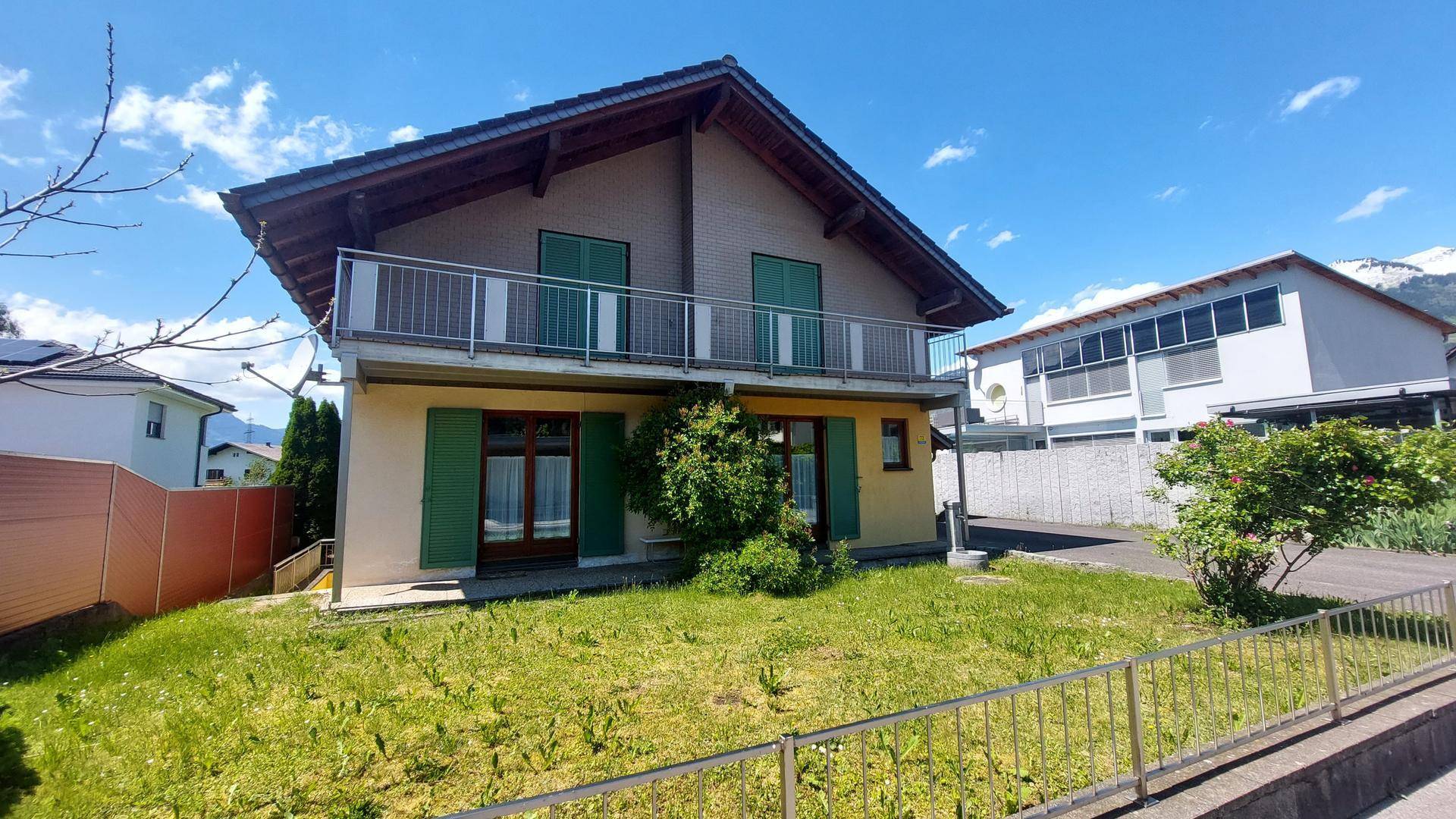 Vorderansicht des Hauses mit Balkon, grünen Fensterläden und gepflegtem Vorgarten an einem sonnigen Tag.