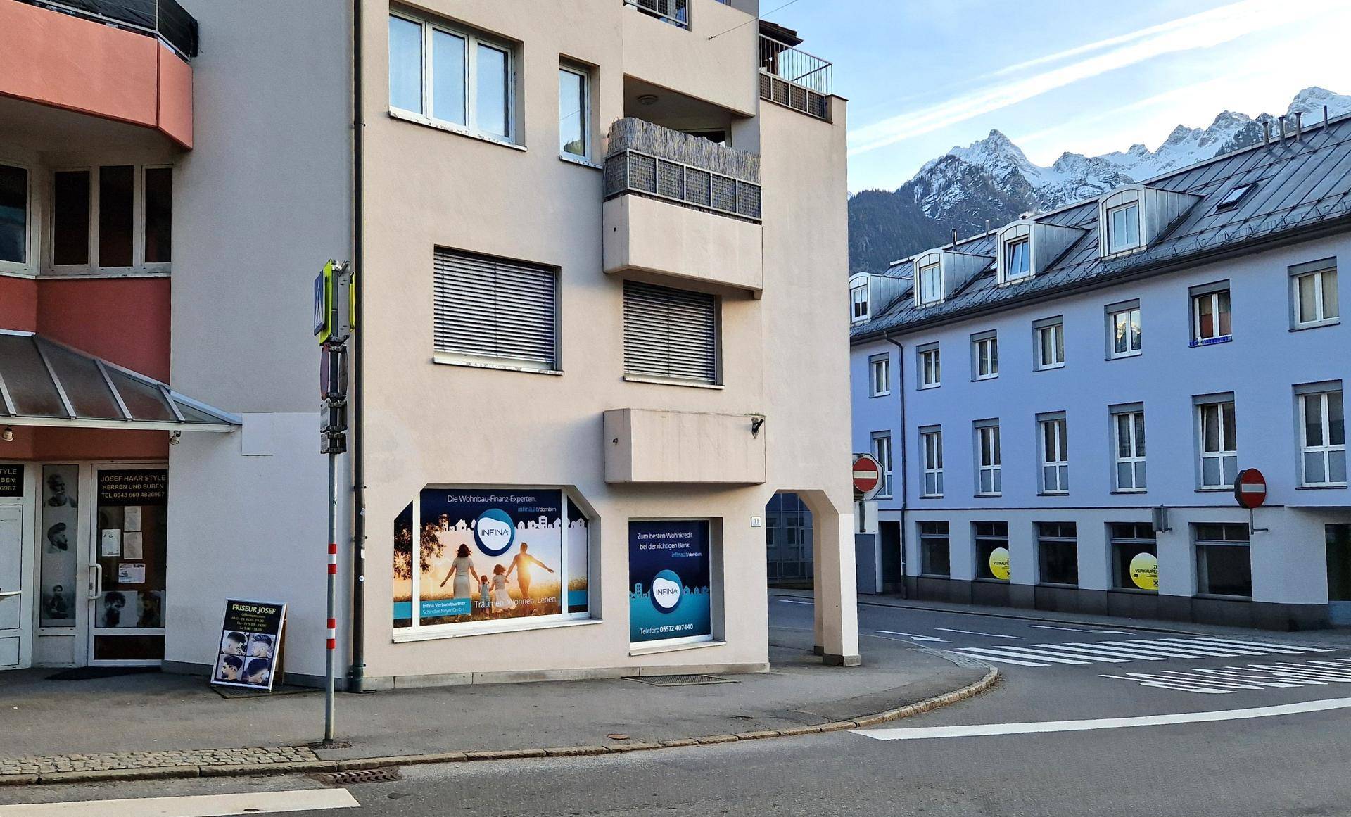 Gebäude an einer Straßenecke mit Schaufenstern und Balkonen, im Hintergrund sind schneebedeckte Berge sichtbar.