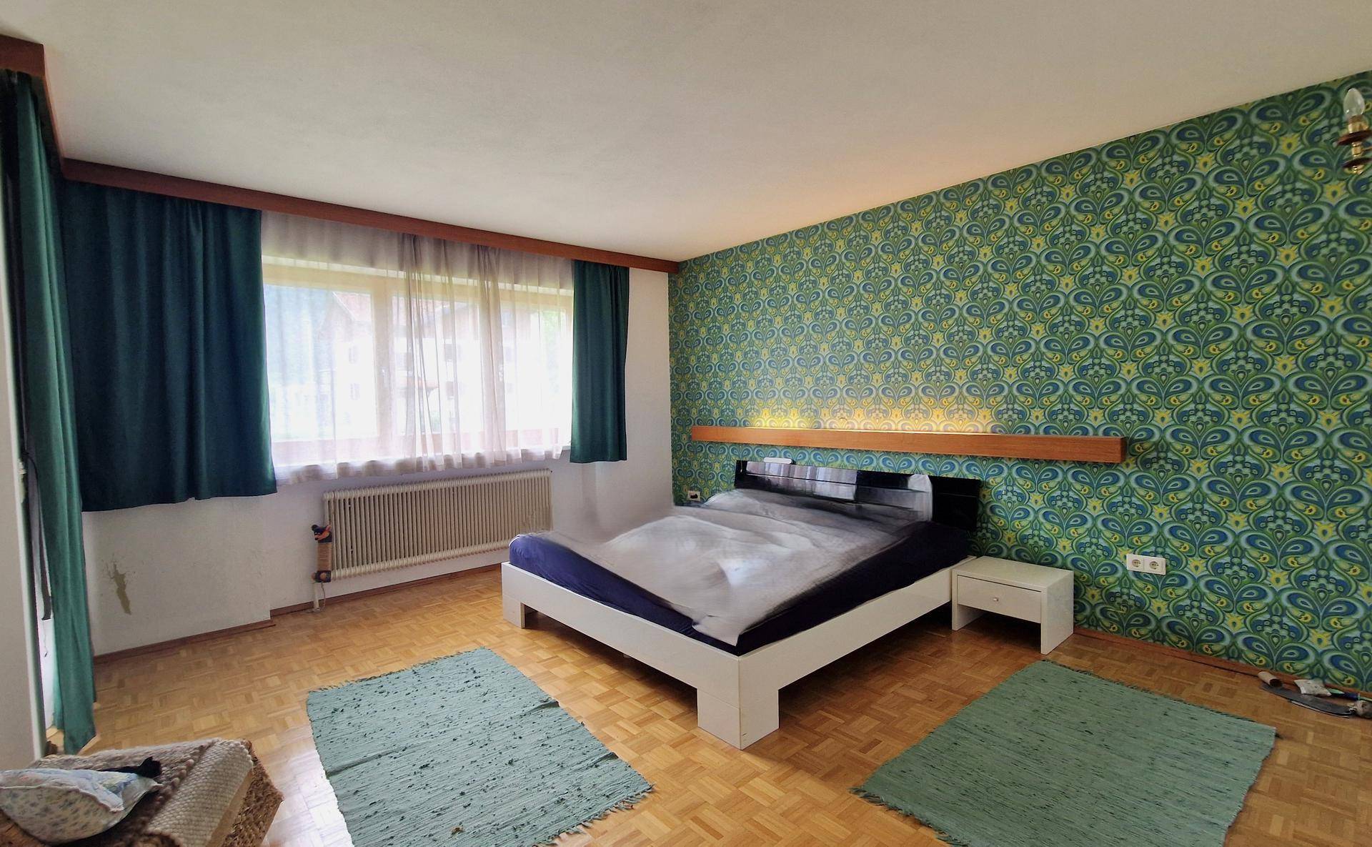 Schlafzimmer mit auffälliger gemusterter Tapete, Parkettboden und großem Fenster.