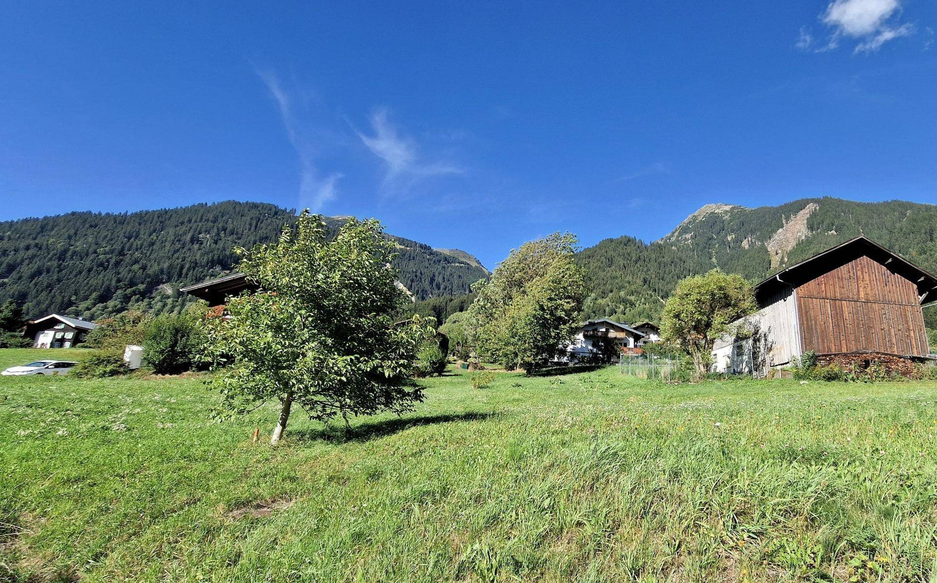 Ländliche Häuser und eine Holzscheune in einer weiten grünen Wiesenlandschaft mit Bergen im Hintergrund.