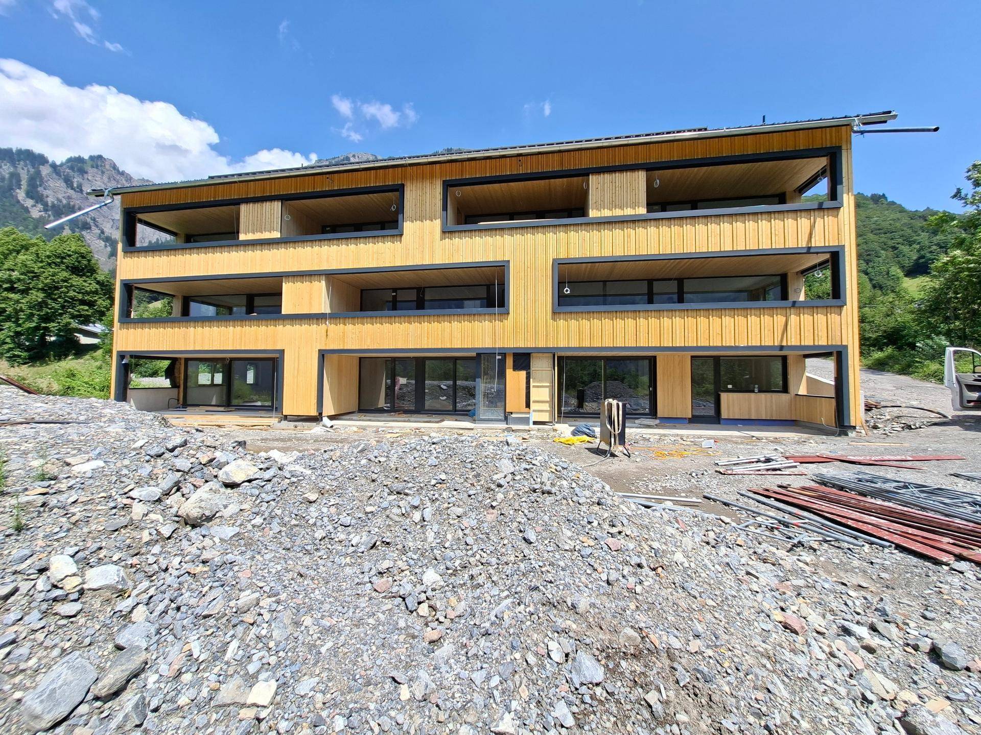 Frontansicht des Neubaus mit Holzfassade, großen Fensterfronten und Bergblick.