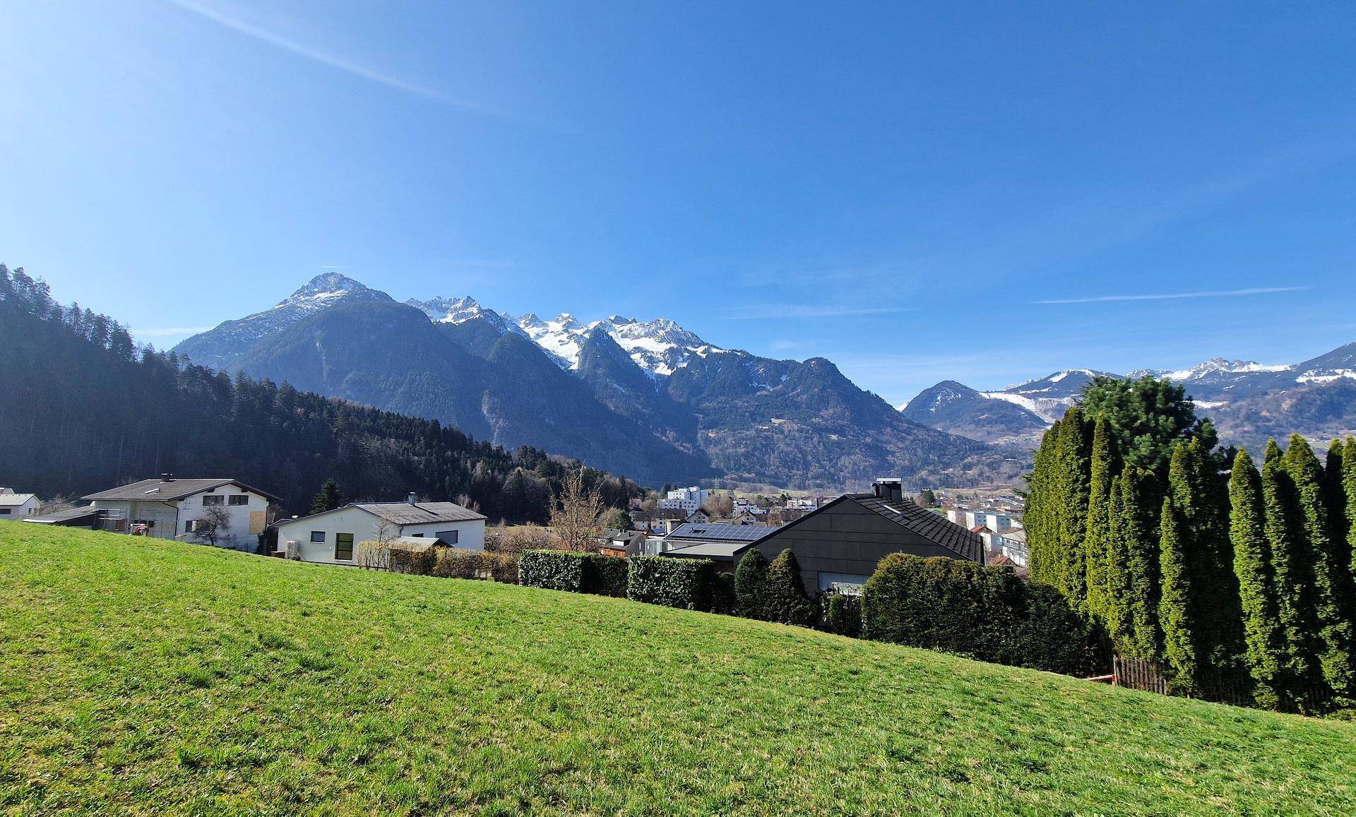 Panoramablick über eine Wohnsiedlung mit grünen Hügeln und schneebedeckten Alpen im Hintergrund.