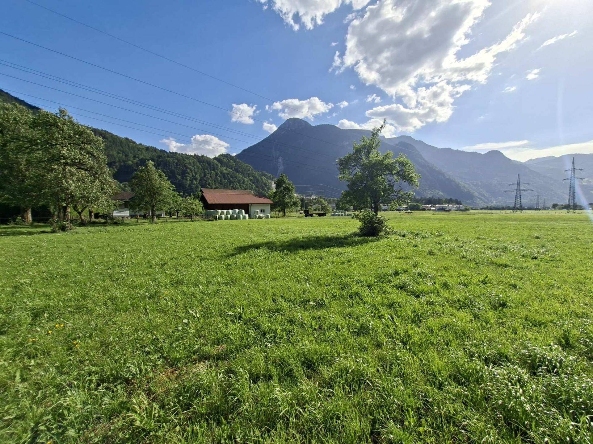 Weite grüne Fläche mit Blick auf die Bergkette und landwirtschaftliche Gebäude in der Ferne.